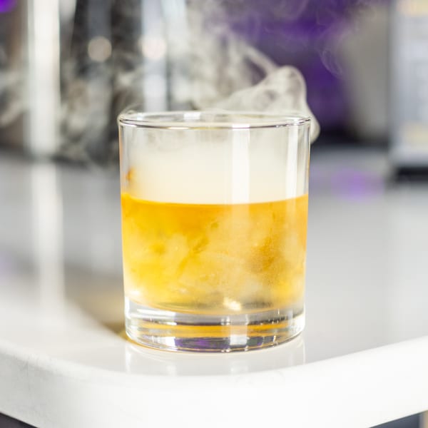 A smoking cocktail in a rocks glass with a golden liquid and white foam, on a white surface.