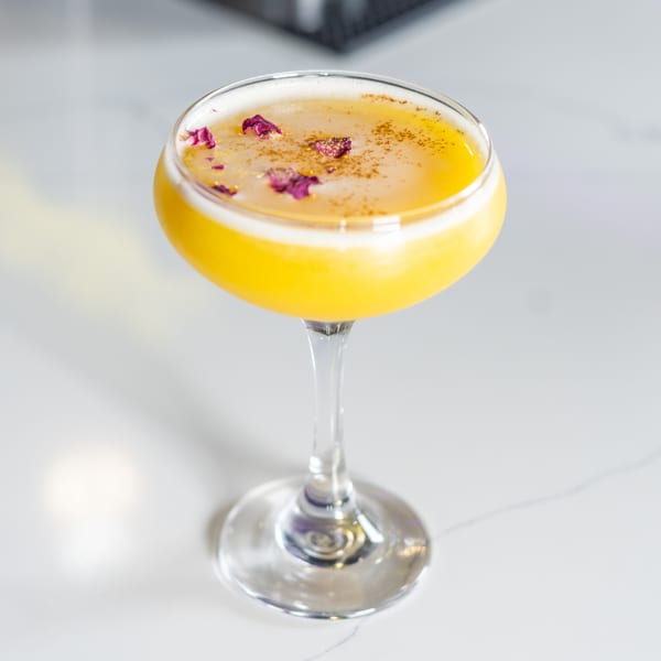 A vibrant yellow cocktail in a coupe glass, garnished with rose petals and spices on a white counter.