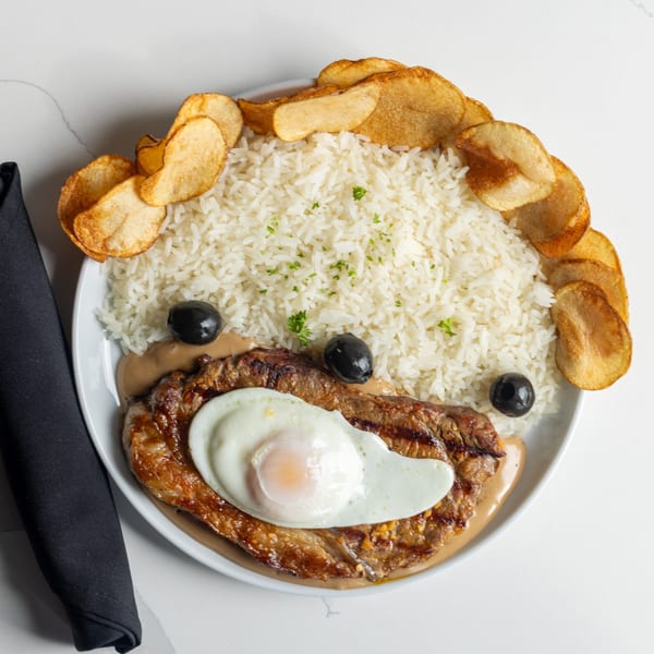 Grilled steak topped with a fried egg, served with rice, potato chips, and olives.