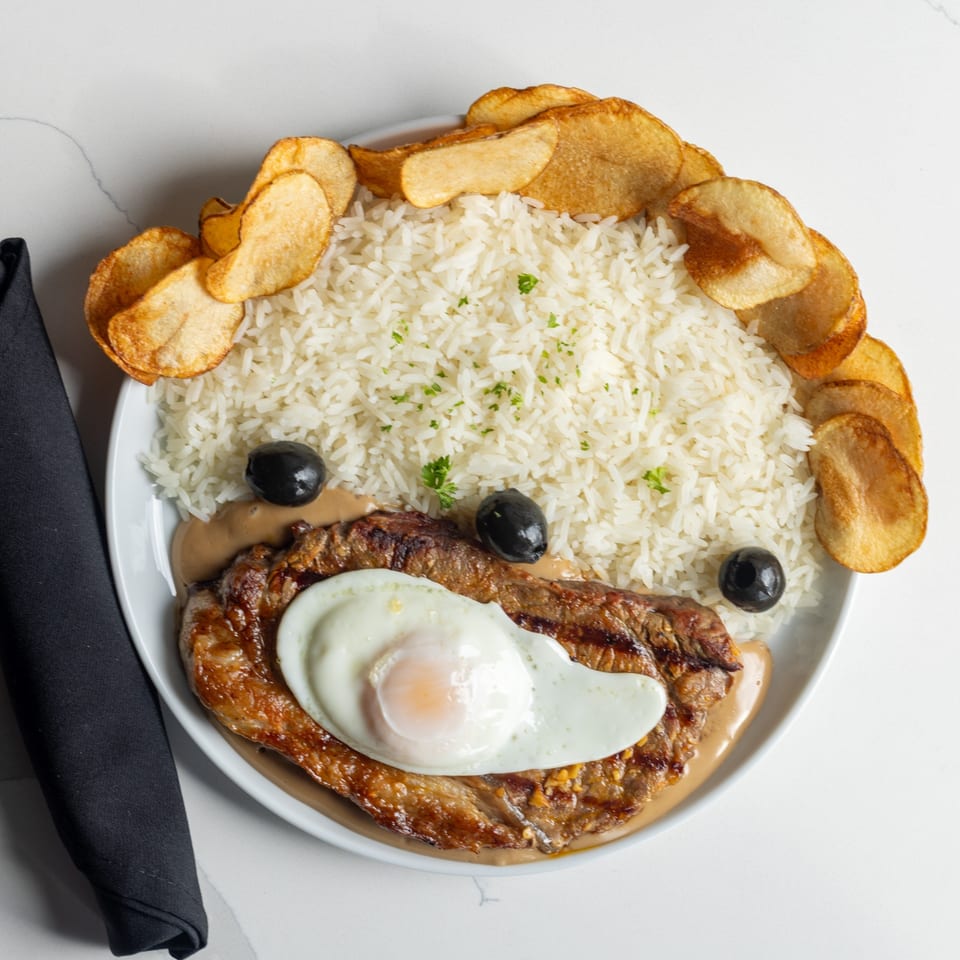 Grilled steak topped with a fried egg, served with rice, potato chips, and olives.
