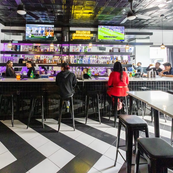 A lively bar scene with patrons seated at a long counter, two bartenders, and TVs showing sports.