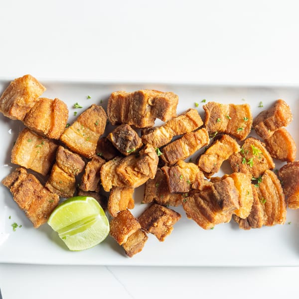 Crispy fried pork belly pieces, chicharrones, garnished with parsley and a lime wedge on a white platter.