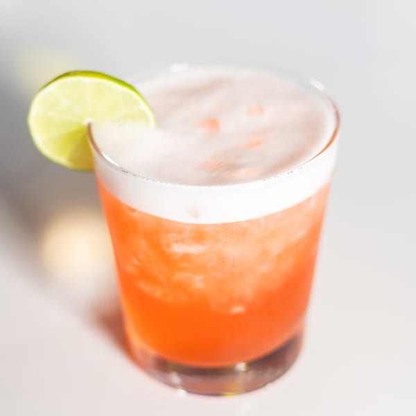 A vibrant orange cocktail with a foamy white top, garnished with a fresh lime slice.