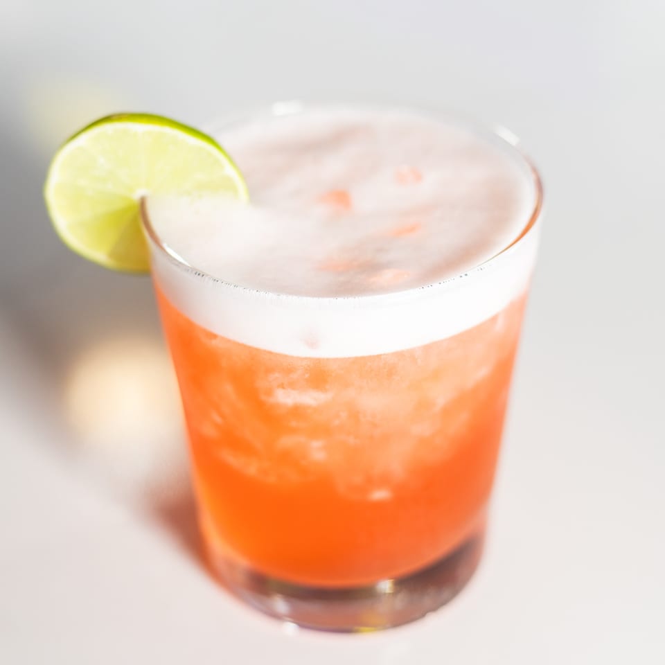A vibrant orange cocktail with a foamy white top, garnished with a fresh lime slice.