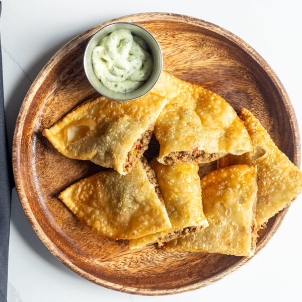 Golden-brown fried empanadas filled with ground meat, served on a wooden plate with a side of green sauce.